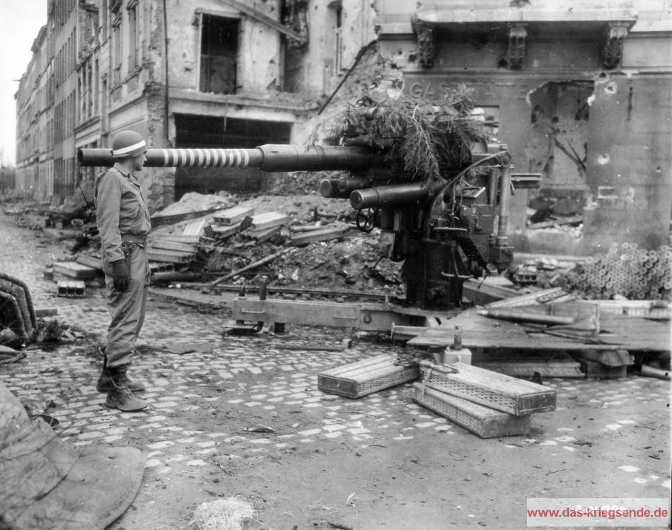Ein amerikanischer Militärpolizist begutachtet eine 88mm FlaK in den mit Trümern übersäten Straßen von Köln. Die Aufnahme wurde durch das Signal Corps der US Army am 7. Mai 1945 gemacht. Die Acht-Acht wurde sehr häufig im Erdkampf gegen Panzer eingesetzt. Die Ringe am Lauf weisen die Abschusszahlen aus. Flak 18 in Köln.
